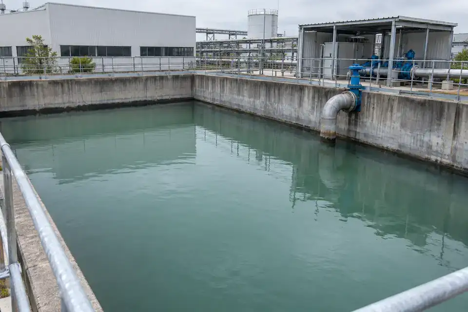 Factory raw-water storage basin with visible water, pipework and an industrial plant in the background